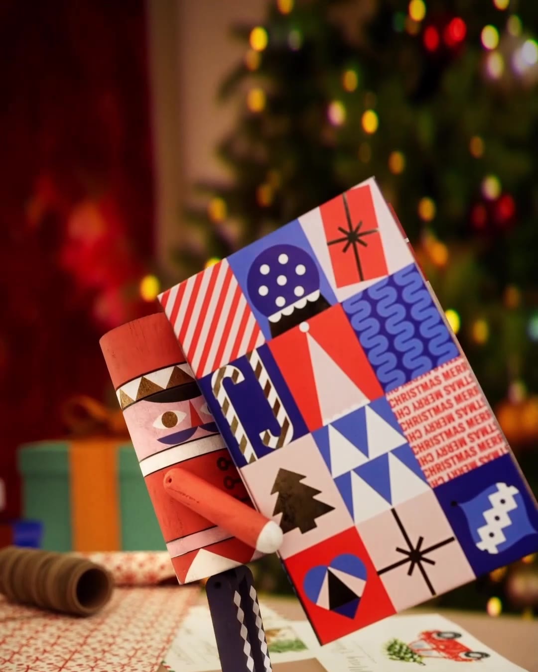 Nötknäpparen bär kalendern över ett bord med presentpapper och kort, framför en julgran med suddiga ljus